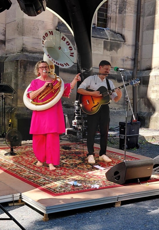 Heike Besen im pinken Hosenanzug am Sousaphon und Julius an der Gitarre auf einer Bühne vor einer Steinkirche.