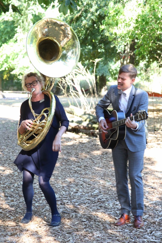 Heike Besen am Sousaphon und Julius im hellen Anzug mit Gitarre bei einer Hochzeit im Grünen.
